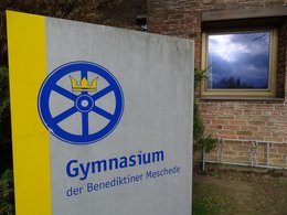 Das Schild vom Benediktiner Gymnasium in Meschede in gelb-blauer Farbe mit Symbol eines Rads mit Krone.