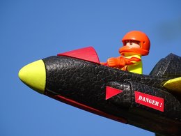 Ein Playmobilmännchen als Flieger mit Helm sitzt in einem Cockpit vor blauem Himmel.