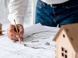 Dies Bild zeigt einen Bauplan für ein Haus, der auf einem Tisch liegt. An dem Bauplan ist eine Person am zeichnen. Links und rechts von dem Bauplan stehen kleine Holzhäuser.