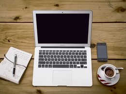 Mobiles Arbeiten zuhause am macbook mit Kaffee. Ein Laptop, ein Glas Wasser, ein Notizbuch und ein Sift auf einem Tisch.