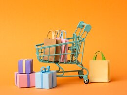 Dieses Bild zeigt bunte Einkaufstaschen, Geschenke die in und neben einem Einkaufswagen stehen. Der Einkaufswagen steht vor einem orangenen Hintergrung.